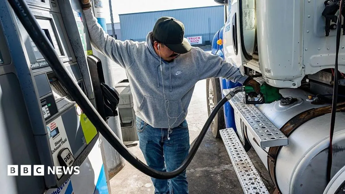 A man wearing a cap and headphones holds a diesel pump against a lorry